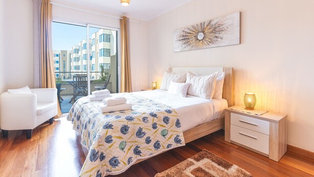 A hotel room with a large bed, a sitting area with a chair, a bedside table with a lamp, and a view of buildings outside.