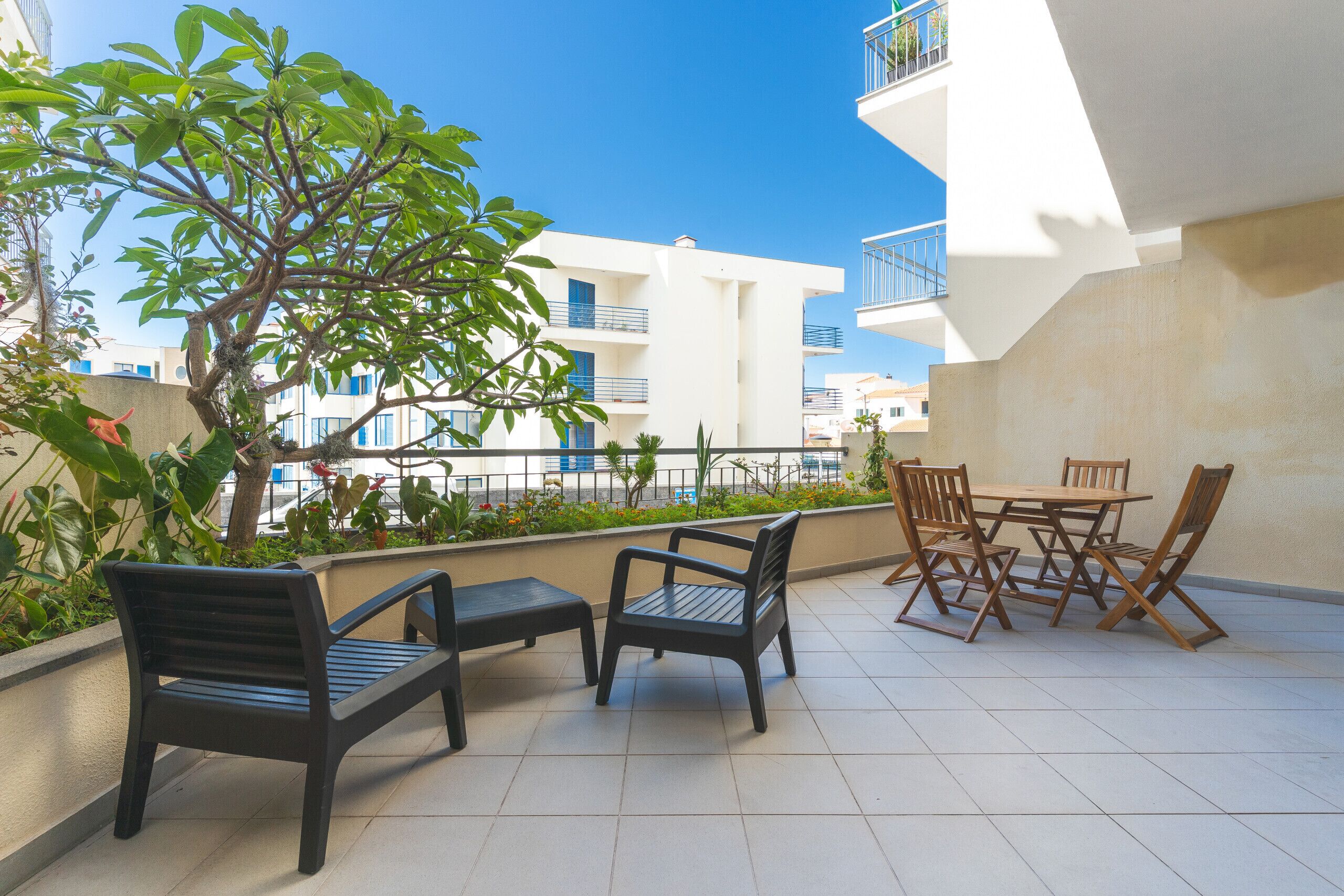 An outdoor seating area with a table and chairs, surrounded by plants and a view of a white building with balconies.