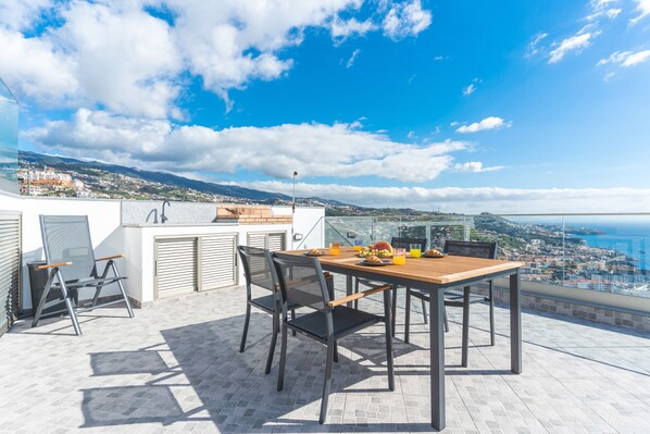 Una terraza con mesa y sillas, vista a la ciudad y al mar, y cielo despejado.