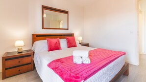 A neatly made bed with a pink blanket and rolled towels, wooden nightstands with lamps, and a mirror on the wall. - Sol Garajau Apartment by Atlantic Holiday (Santa Cruz)