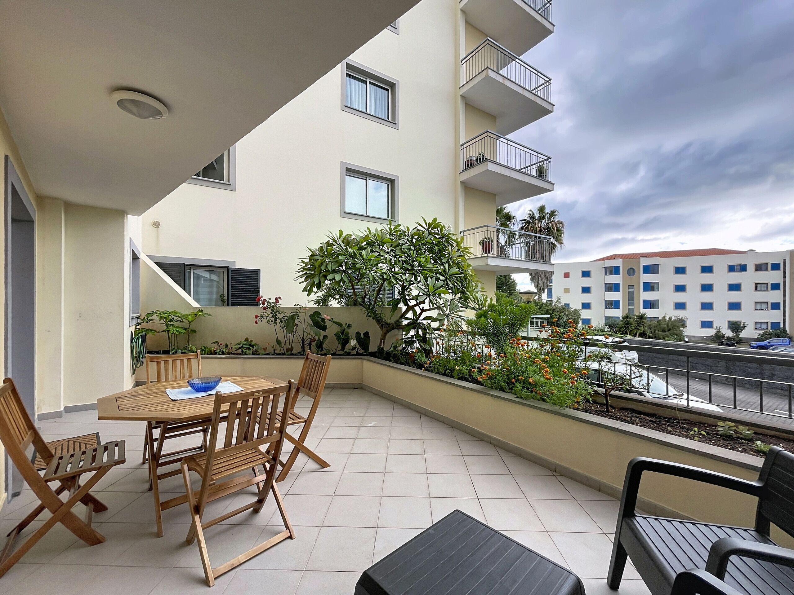 A balcony with a wooden table and chairs, surrounded by potted plants and flowers.