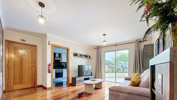 A modern living room with a wooden floor, a sofa, a coffee table, a TV, and a sliding glass door leading to a balcony. - Judite Home Apartment by Atlantic Holiday (Santa Cruz)