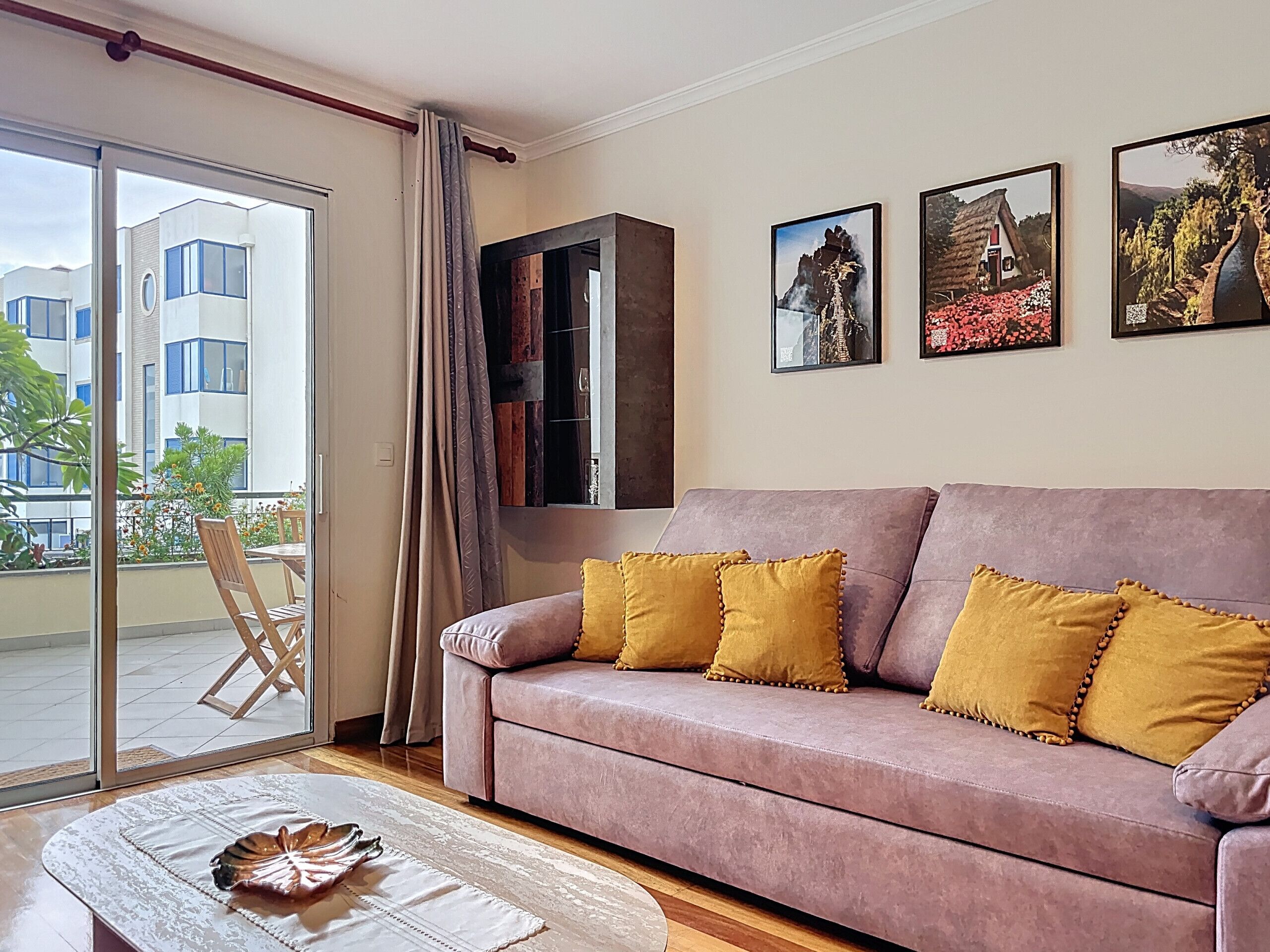 A living room with a pink sofa, yellow cushions, a glass door leading to a balcony, and three framed pictures on the wall.