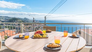 Ein runder Holztisch mit Frühstücksangeboten, darunter Croissants, Obst und Saft, mit Blick auf die Küste.