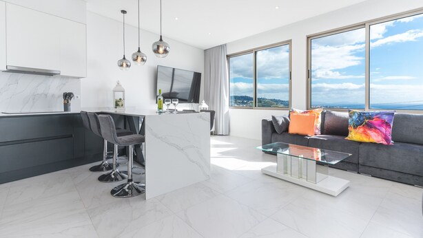 Eine moderne Küche mit Marmorarbeitsplatte, Barhocker und einer großen Kochinsel. Ein Essbereich mit einem Glastisch und einem grauen Sofa mit bunten Kissen. Ein großes Fenster bietet Blick auf die Stadt.