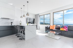 Una cocina moderna con encimera de mármol, taburetes de bar y una isla grande. Hay un comedor con una mesa de centro de vidrio y un sofá gris con cojines de colores. Un ventanal ofrece vistas a la ciudad.