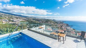Ein Pool auf dem Dach mit Glasbrüstung, einem Holztisch und einem Stuhl, mit Blick auf eine Küstenstadt mit Gebäuden und dem Meer.
