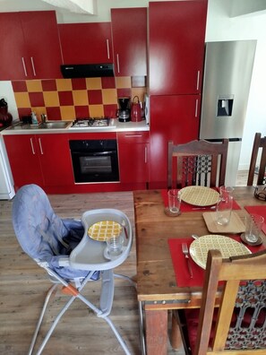A modern kitchen with red cabinets, a stainless steel refrigerator, and a dining area with a wooden table set for two. - Chez Béa*** Vacation Rental: fenced-in yard, dogs welcome, quiet, all-inclusive rate (Pleumartin)