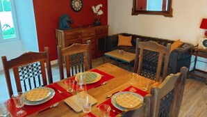 Un'area pranzo con un tavolo di legno apparecchiato per quattro persone, circondato da sedie con decorazioni elaborate. Un sottopiatti rosso ricopre il tavolo, sul quale sono presenti calici da vino e piatti. A dietro il tavolo, sulla parete, si trova un orologio e una pianta in vaso.