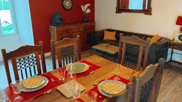 Een eethoek met een houten tafel voor vier personen, omringd door stoelen met gedetailleerde rugleuningen. Een rode placemat bedekt de tafel, met wijnglazen en borden erop. Op de muur achter de tafel staan een klok en een potplant.