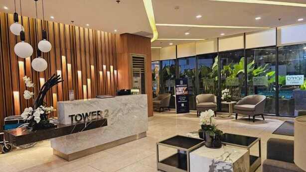 Un moderno vestíbulo de hotel con una recepción identificada como "TOWER 2", zona de estar y grandes ventanales con plantas.