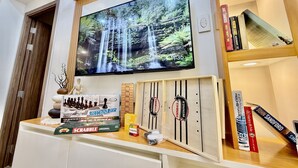 Una sala de estar con un televisor de pantalla plana montado en la pared, un estante de madera con libros y juegos de mesa, y una mesita con adornos.