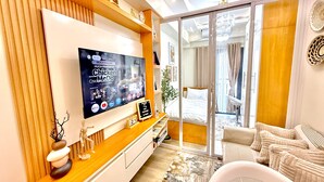 Habitación de hotel moderna con un televisor de pantalla plana grande montado en una pared de madera, una zona de estar acogedora con un sofá y sillones, una mesita con una planta y una puerta de cristal corredera que da a un balcón.