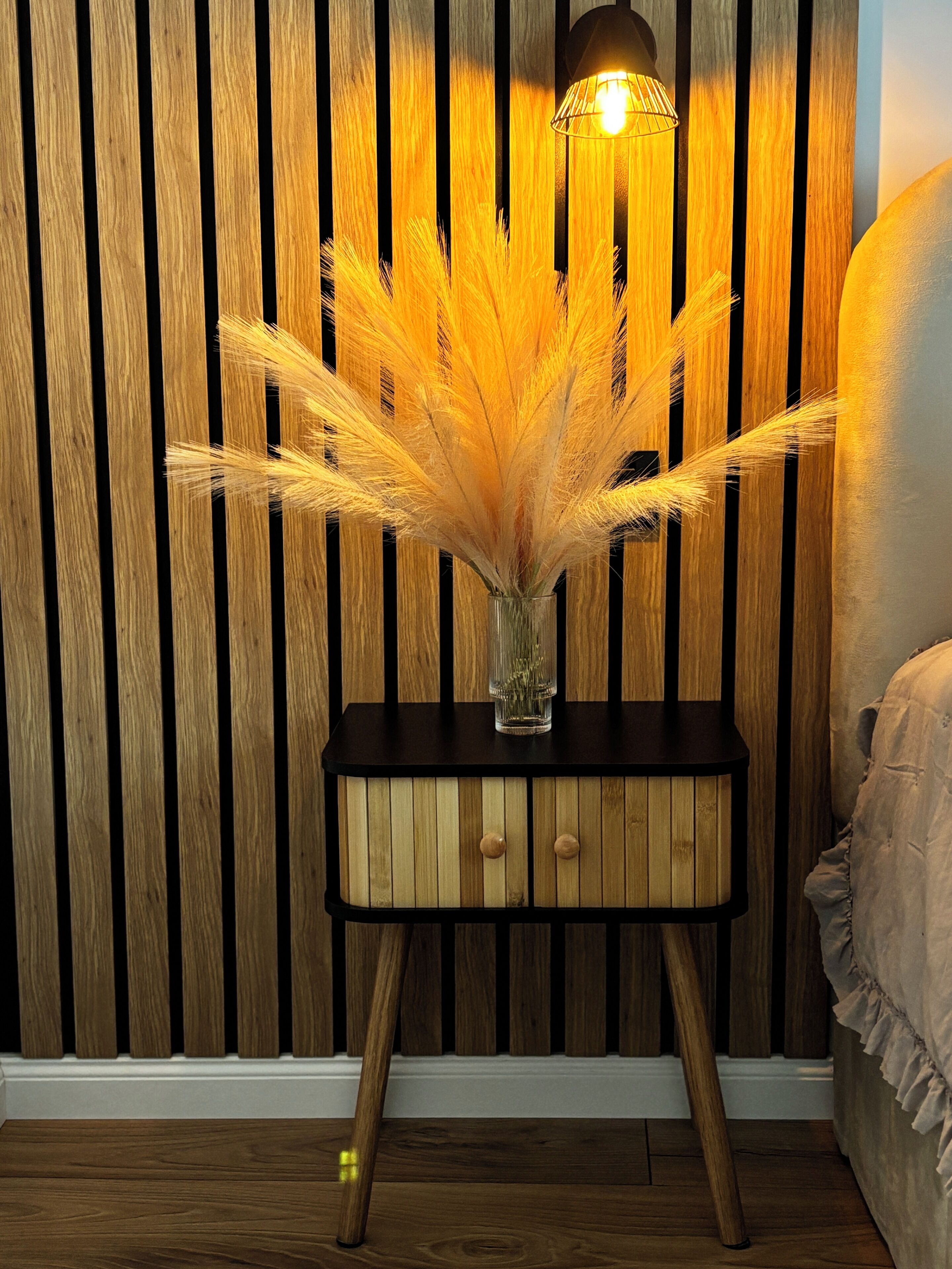 A bedside table with a vase of dried pampas grass and a lamp.