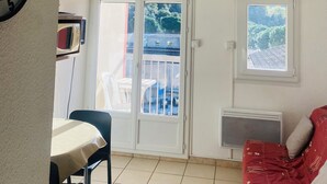 Une pièce avec une horloge, une table entourée de chaises, un canapé rouge, une porte-fenêtre coulissante et une fenêtre donnant sur des arbres.