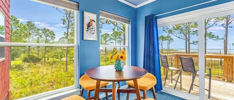 A room with a large window overlooking a deck and trees, a round table with chairs, and a framed picture on the wall.
