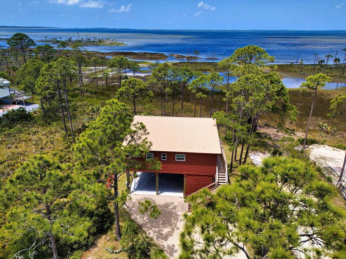 A single-story house with a red exterior and a metal roof, surrounded by pine trees and a body of water in the background.