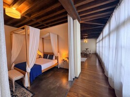 Una cama con dosel adornada con cortinas blancas, suelo de madera y un área de comedor al fondo.