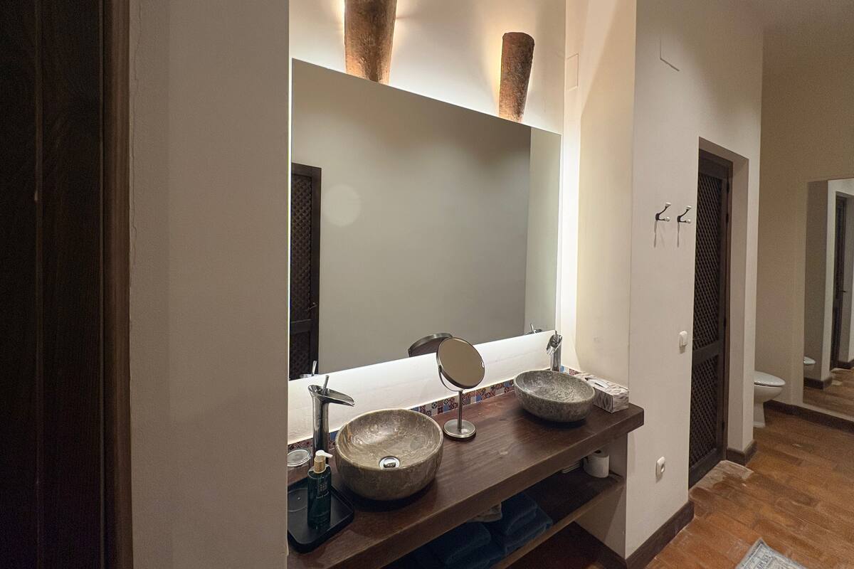 A bathroom with a large mirror, two stone sinks, a handheld mirror, and a towel rack.