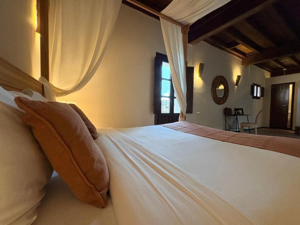 A bedroom with a bed, a window, a mirror, and a small table with a chair.