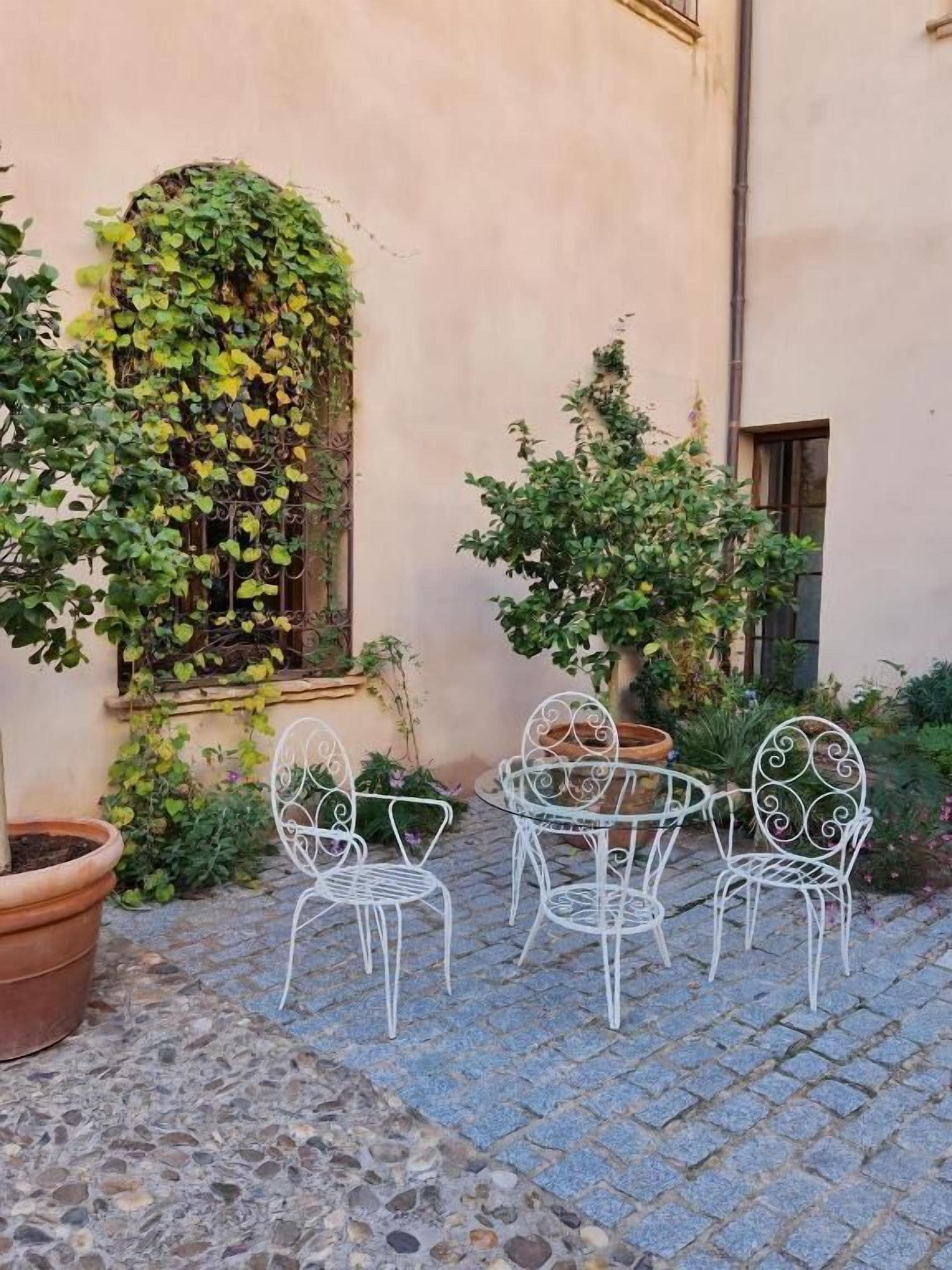 A courtyard with a stone floor, white metal chairs, and potted plants.