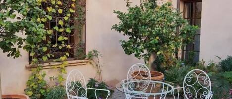 A courtyard with a stone floor, white metal chairs, and potted plants.