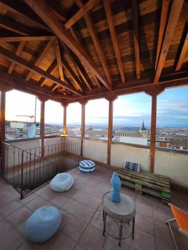 A covered outdoor area with a view of a cityscape, featuring a wooden ceiling, a metal railing, and various seating options. - Casa del Armiño (Toledo)