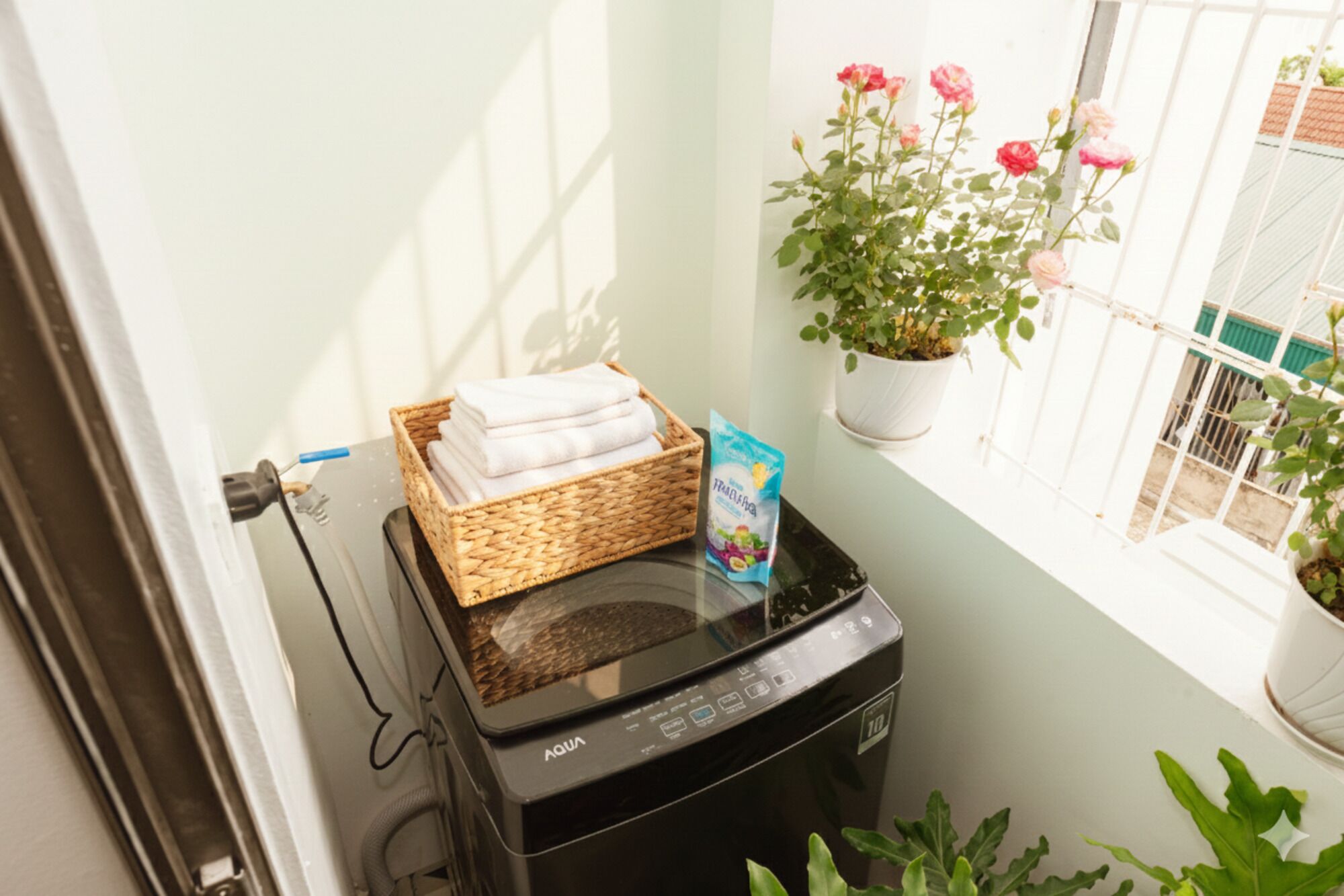 Une petite pièce équipée d’un lave-linge, d’un panier de serviettes pliées, d’une plante en pot aux fleurs roses et d’une fenêtre donnant sur un immeuble.