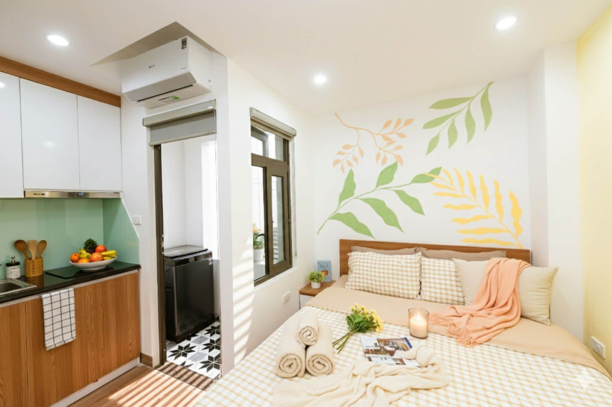 A compact hotel room with a kitchenette, a bed with a checkered bedspread, and a wall with a leaf mural.