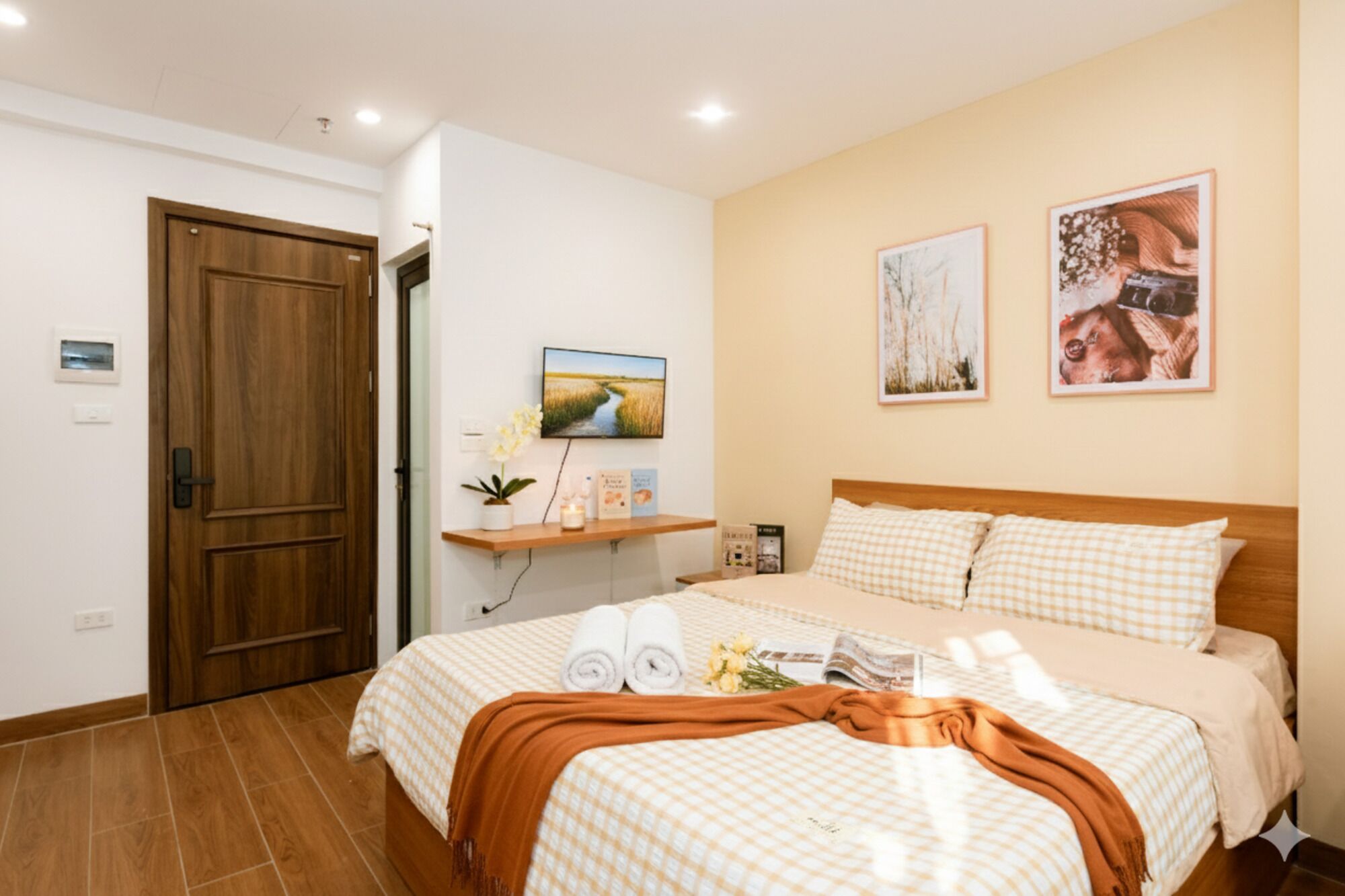 A hotel room with a wooden door, a bed with checkered bedding, a bedside table with a lamp, and two framed pictures on the wall.