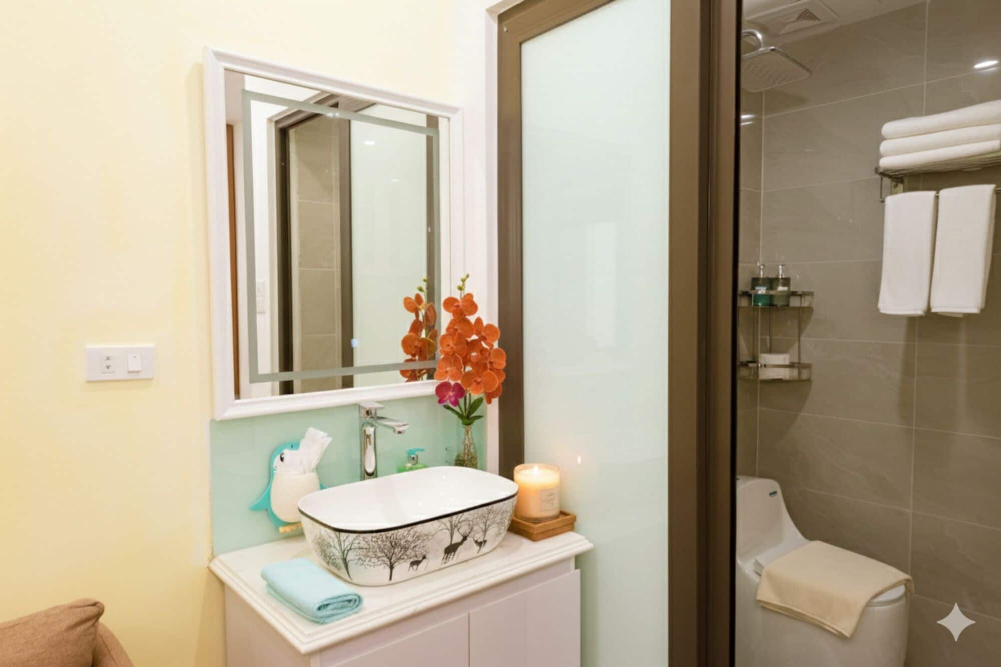 A bathroom with a white sink, a mirror, a vase with flowers, a candle, and a glass shower enclosure.