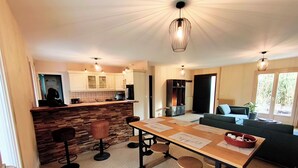 Een moderne keuken met een centraal eiland, barstoelen en een eettafel met papieren. Er is een zithoek met een bank en een potplant.