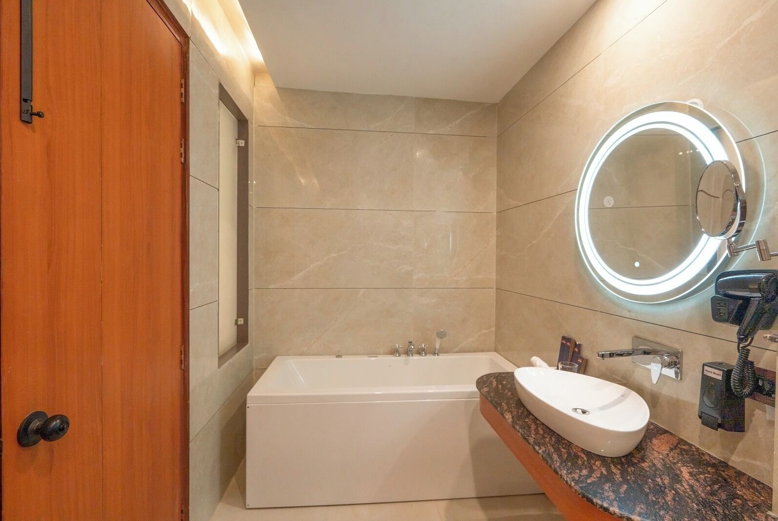Ein modernes Badezimmer mit einer großzügigen Badewanne, einem runden Spiegel mit Beleuchtung, einer Marmorplatte und einer Holztür.