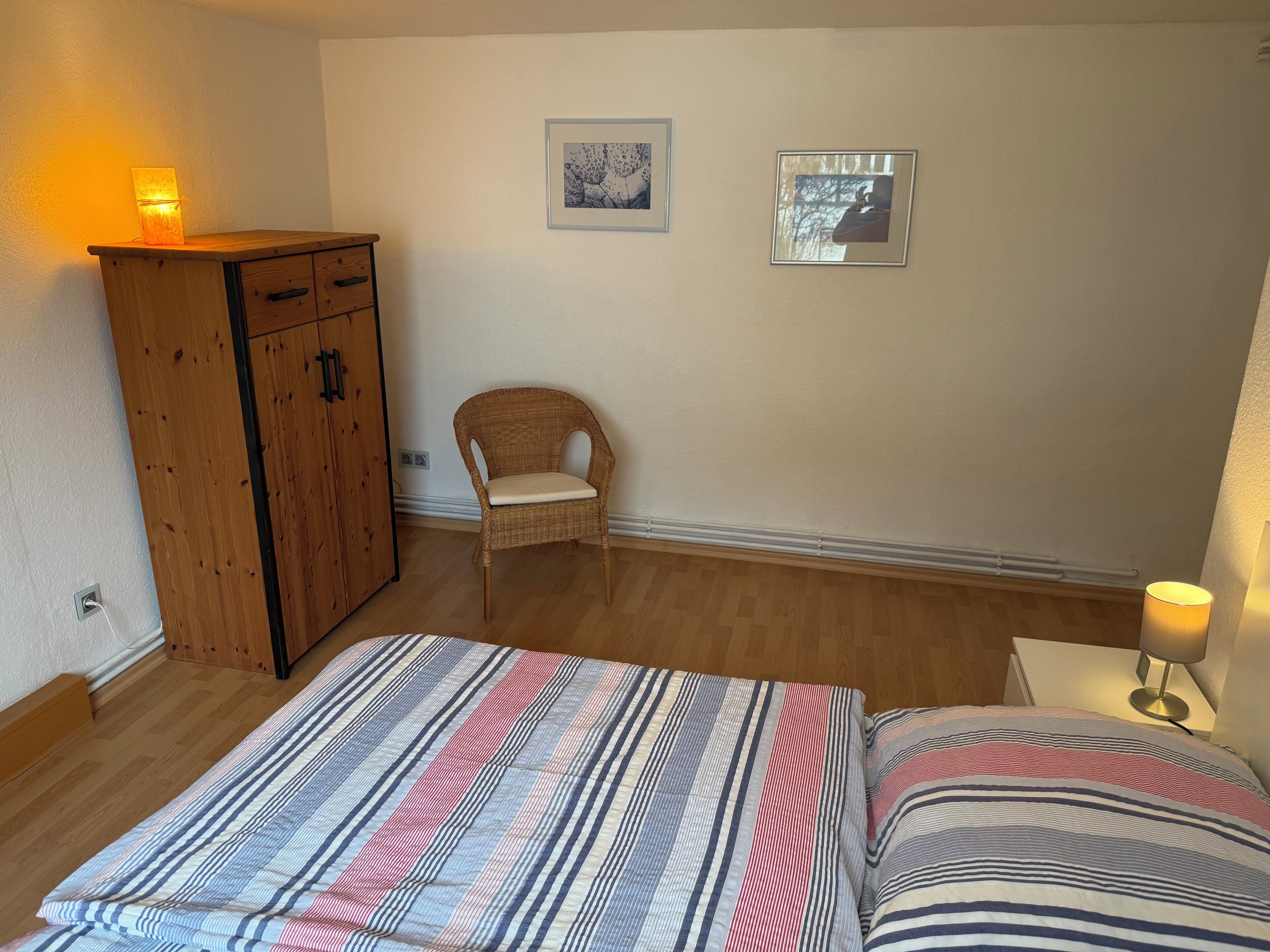 Une chambre à coucher avec un lit, une armoire en bois, une chaise en osier et deux tableaux encadrés accrochés au mur.