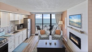A modern living room with a kitchen, a fireplace, and a balcony with a view of the ocean. - Beach Colony Oceanfront w/ Indoor & Outdoor Pools (Myrtle Beach)
