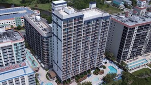 A high-rise hotel with multiple balconies, a rooftop pool, and a landscaped pool area with lounge chairs. - Beach Colony Oceanfront w/ Indoor & Outdoor Pools (Myrtle Beach)