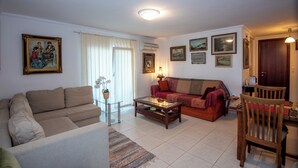 A living room with a sofa, a coffee table, a wooden chair, and a dining table. There are framed pictures on the wall and a potted plant. - Gigi Star Tatiana Luxury Apartment (Zakynthos)