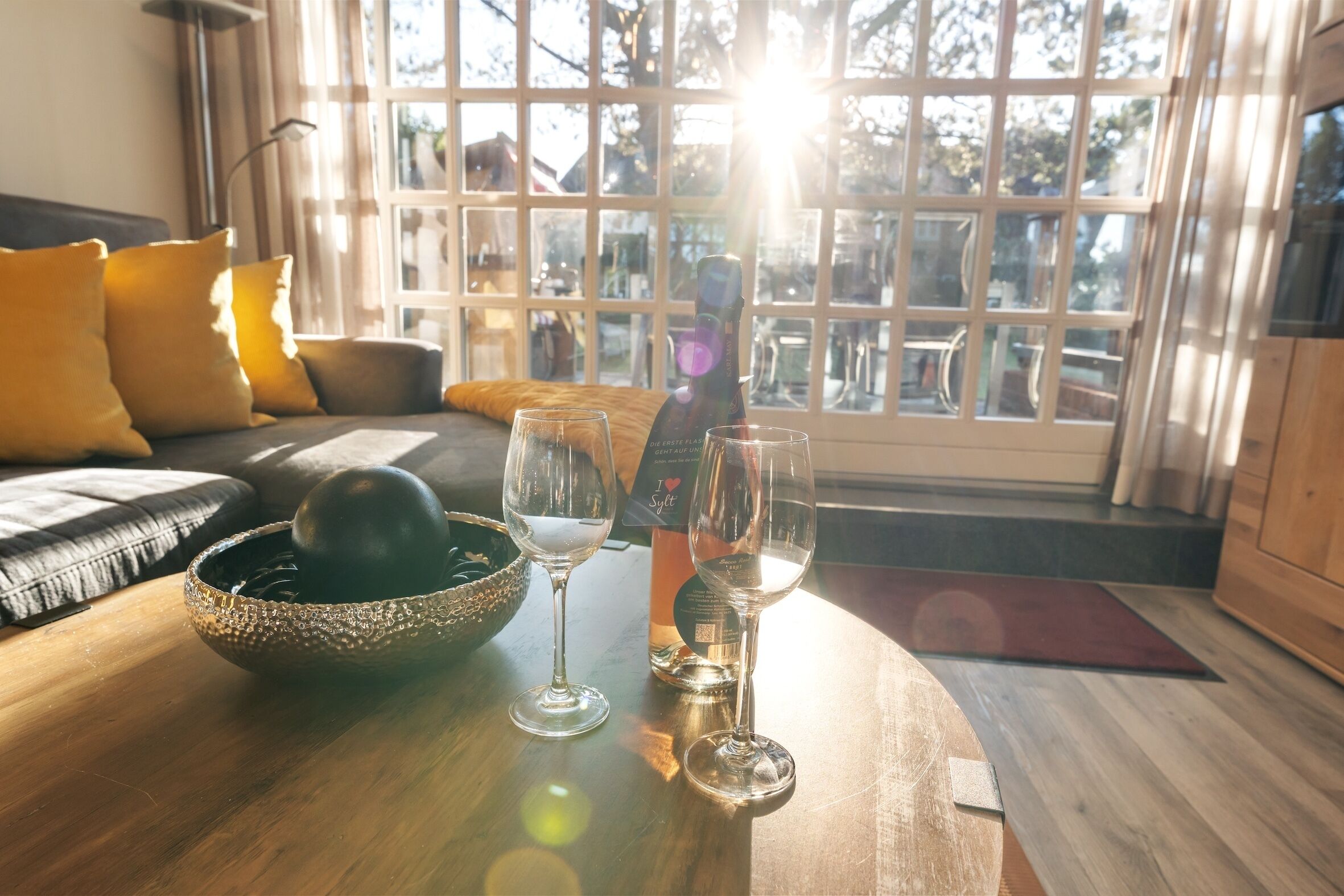 A wooden table with a decorative bowl, two wine glasses, and a bottle of wine in a room with a large window, a sofa with cushions, and a lamp.