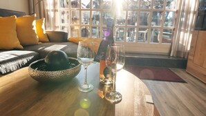 A wooden table with a decorative bowl, two wine glasses, and a bottle of wine in a room with a large window, a sofa with cushions, and a lamp. - Vacation home "Hüs Swarte Wall" (Kampen (Sylt))