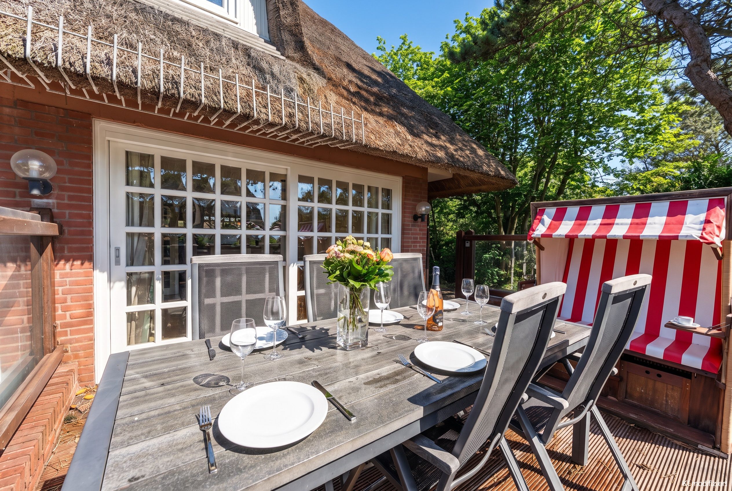 Een terras met een eettafel gedekt voor twee personen, een rieten dak en een rood-wit gestreept terrasoverkapping.