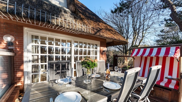 A patio with a table set for dining, a thatched roof, and a striped awning. - Vacation home "Hüs Swarte Wall" (Kampen (Sylt))