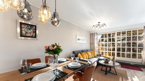 A dining area with a wooden table set for four, surrounded by modern chairs. There is a sofa with yellow cushions in the background, and a large window with curtains. - Vacation home "Hüs Swarte Wall" (Kampen (Sylt))