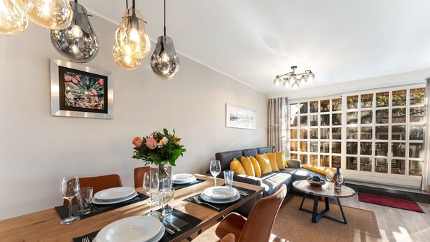 A dining area with a wooden table set for four, surrounded by modern chairs. There is a sofa with yellow cushions in the background, and a large window with curtains. - Vacation home "Hüs Swarte Wall" (Kampen (Sylt))