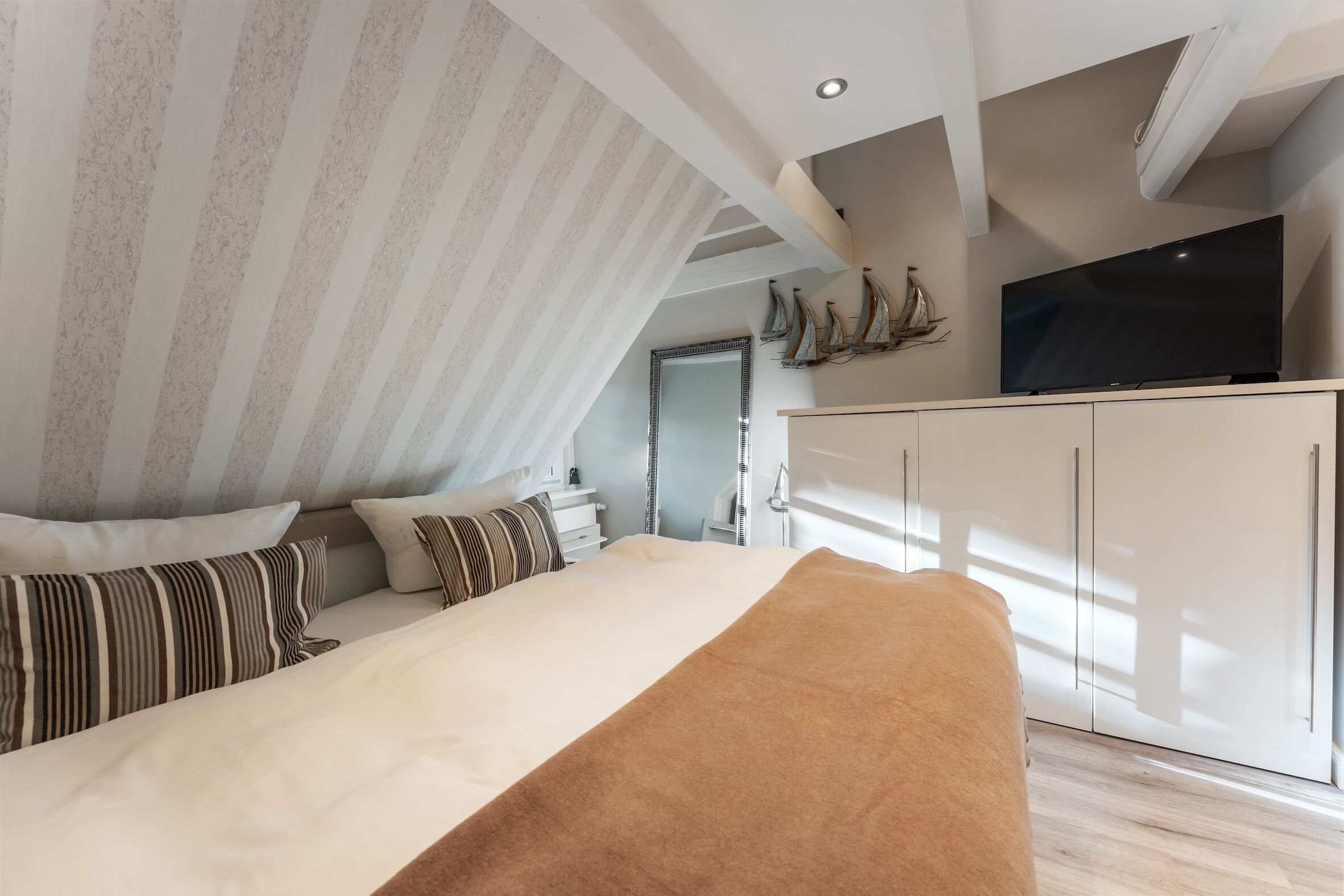 Une chambre à coucher avec un lit, des oreillers, un téléviseur posé sur un meuble et un plafond avec des poutres apparentes.