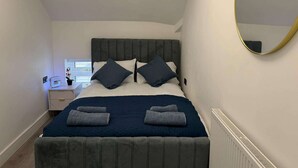 A bedroom with a bed, pillows, a bedside table, a mirror, and a radiator. - Cardiff Central Gem - 2 Bed Apartment (Cardiff)