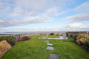 A grassy outdoor area with a stone wall, a wooden bench, and a paved path leading to a wooden deck with chairs. - Host Stay The Shipshape (Seahouses)