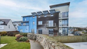 Un immeuble résidentiel moderne à plusieurs étages, doté de balcons, d’un mur de soutènement en pierre et d’une zone herbeuse au premier plan.