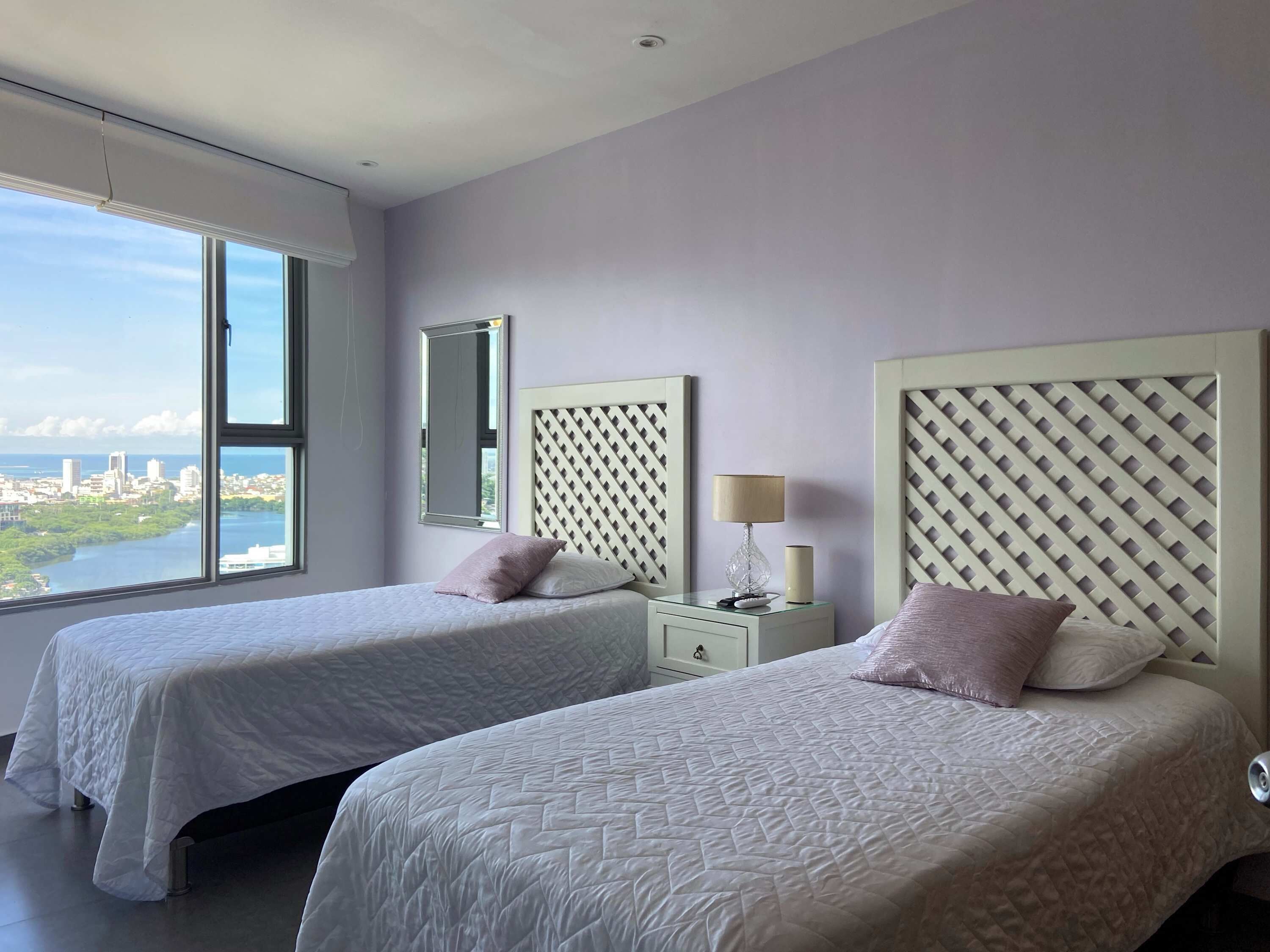 A hotel room with two beds, a nightstand, a lamp, and a large window offering a city view.