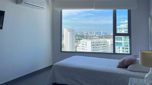 A hotel room with a large window offering a city view, a bed with white bedding, a bedside table with a lamp, and a remote control. - w Amazing 3BR w Ocean View in Marbella (Cartagena)