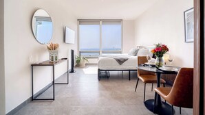 Een moderne hotelkamer met een bed, een eettafel met stoelen, een klein bijzettafeltje, een groot raam en een spiegel aan de muur.